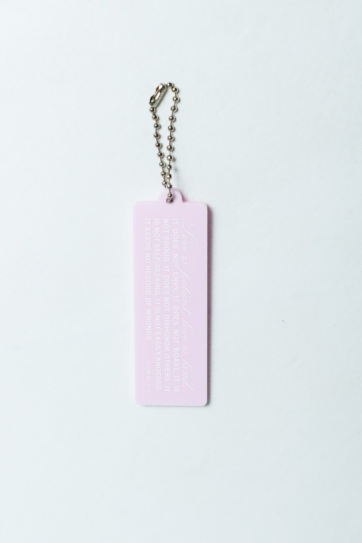 A light pink rectangular Christian acrylic keychain/charm with embossed text – Bible verse 1 Corinthians 13:4-5, attached to a silver ball chain, shown against a plain white background.