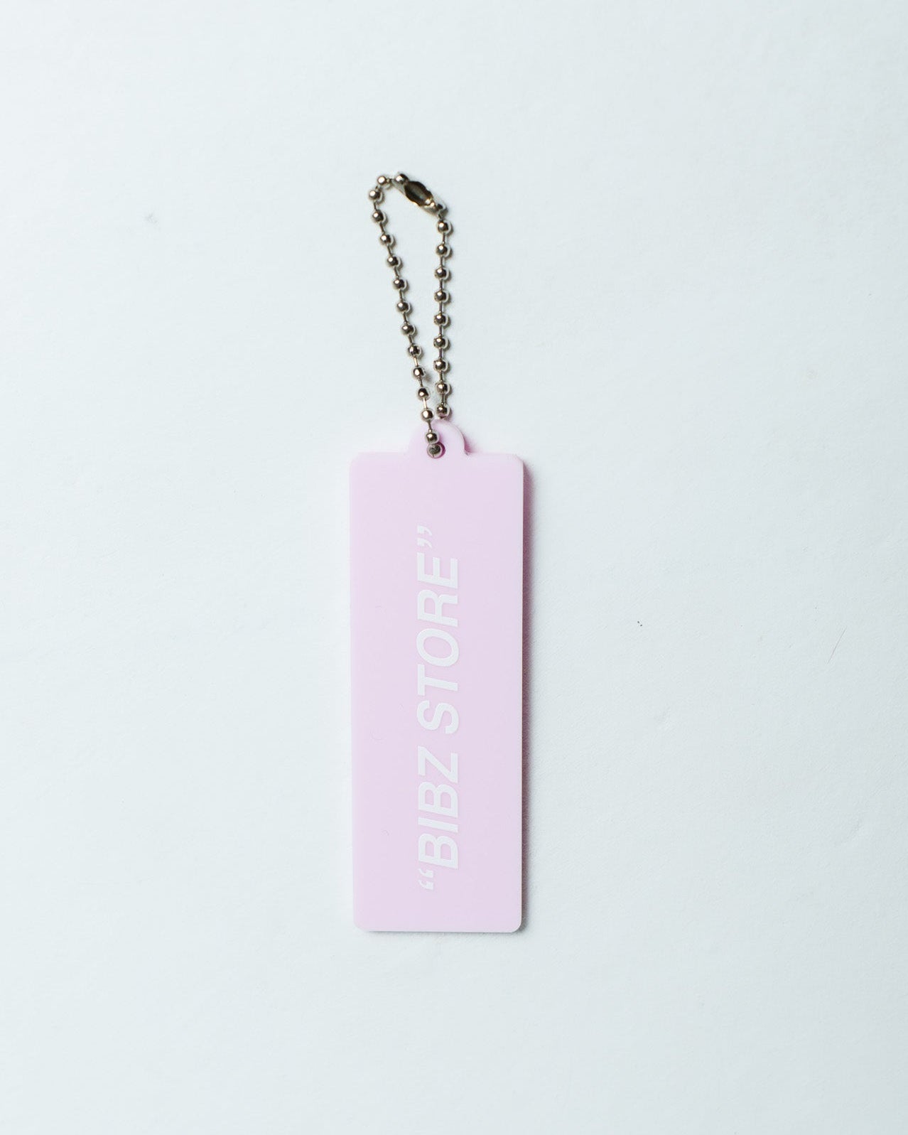 A light pink rectangular keychain tag with the text "BIBZ STORE" printed vertically in white, attached to a silver ball chain, on a plain white background.