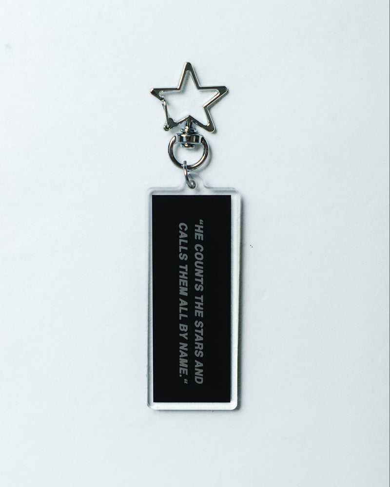 A black rectangular acrylic keychain with white text reading 'HE COUNTS THE STARS AND CALLS THEM ALL BY NAME' and a silver metal star keyring, shown on a plain white background.